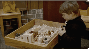 A young boy stands next to a wooden table, looking at a miniature castle model constructed with various small pieces, set inside a wooden box.