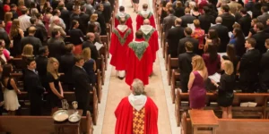 Une procession religieuse avec des membres du clergé vêtus de robes rouges et vertes marchant dans l'allée centrale, entourés de fidèles assis et debout.