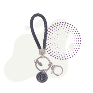 The Charm Key Fob features a black braided keychain adorned with a circular silver charm and a lobster clasp, showcased against a white background with abstract purple and light gray shapes.