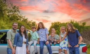 A family of seven poses by a white truck at sunset, with greenery in the background and colorful clouds in the sky.