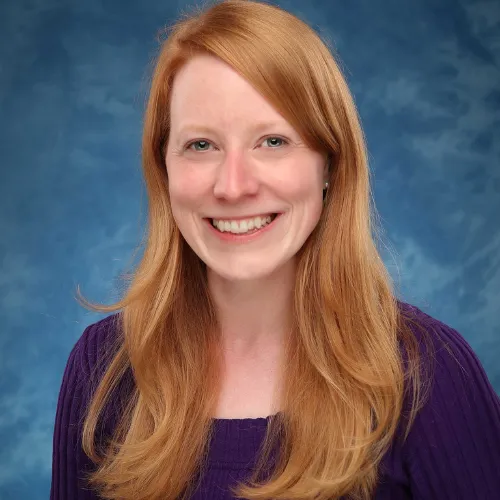 A woman with long red hair and fair skin smiles at the camera, wearing a purple top in front of a blue, cloudy background.