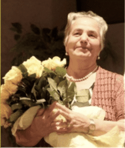 An older woman with gray hair holds a large bouquet of yellow roses and smiles. She wears a pearl necklace and a textured pink cardigan.