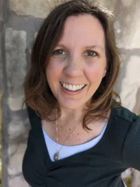 A woman with brown hair, wearing a dark green top and a necklace, stands in front of a stone wall and smiles at the camera.