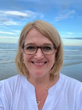 A woman with blonde hair and glasses smiles at the camera while standing on a beach with the ocean and blue sky in the background.