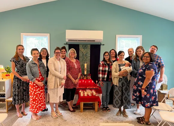 A group of people pose indoors around a table with candles and a large white candle, in a room with teal walls and two windows.