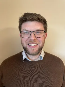 A man with short brown hair, glasses, and a beard smiles at the camera. He is wearing a brown sweater over a collared shirt, standing in front of a beige wall.