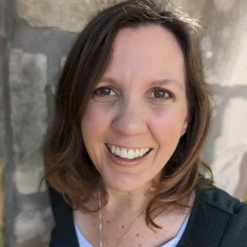 Woman with shoulder-length brown hair smiles at the camera, standing in front of a stone wall.