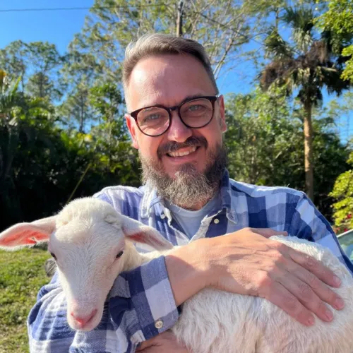 A man wearing glasses and a plaid shirt is smiling and holding a white lamb outdoors on a sunny day.