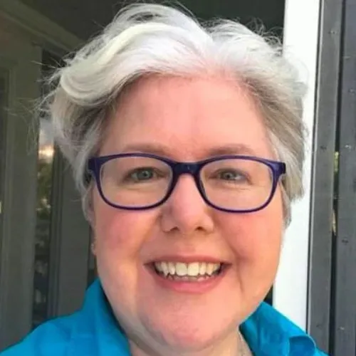 A person with short gray hair and glasses smiles at the camera while standing outside near a door.