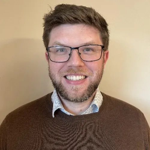 A man with short brown hair, glasses, and a beard is smiling at the camera. He is wearing a brown sweater over a white collared shirt. The background is plain beige.