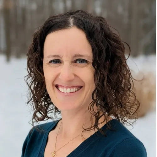 Woman with curly brown hair and a blue top smiles outdoors in a snowy, wooded area.