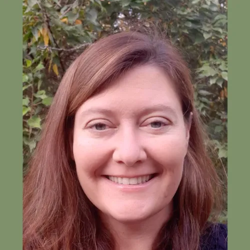 A woman with long brown hair smiles at the camera, standing outdoors in front of green foliage.