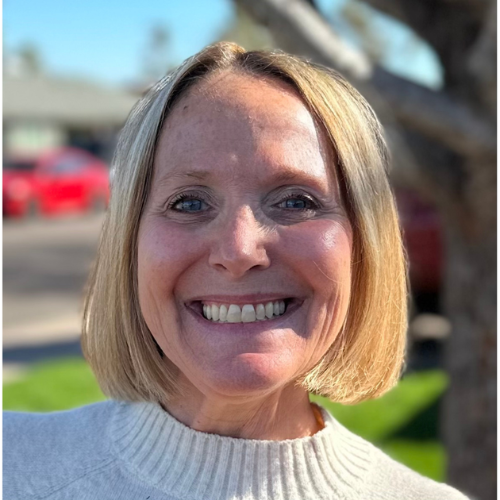 A woman with straight blonde hair and a cream sweater smiles outdoors, with blurred trees and cars in the background.