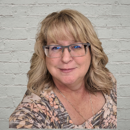 A middle-aged woman with blonde hair and glasses smiles at the camera, standing in front of a light gray brick wall. She is wearing a patterned top and a necklace.