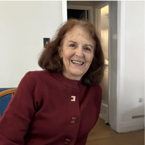 An older woman with shoulder-length brown hair, wearing a maroon jacket, sits indoors and smiles at the camera.