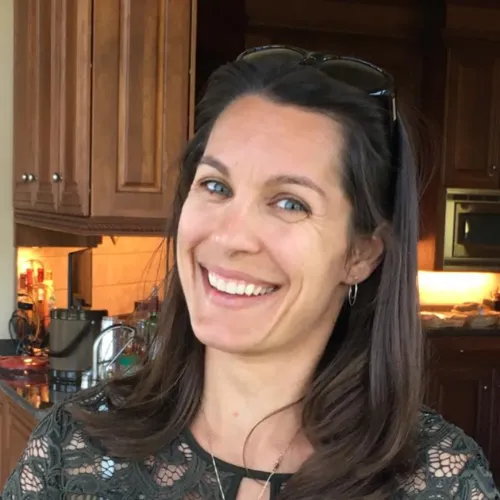 A woman with long brown hair and blue eyes smiles in a kitchen with wooden cabinets and various appliances in the background.