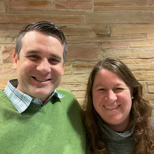 Two people smiling and posing together in front of a tan brick wall. The person on the left wears a green sweater; the person on the right has long brown hair and a gray top.