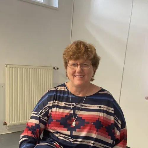 A smiling woman with short brown hair and glasses sits indoors, wearing a blue, red, and white patterned top. A radiator and white wall are visible in the background.