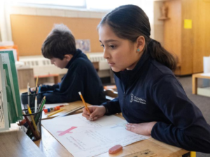Two elementary school students sit at desks working on drawings and writing assignments in a classroom setting.